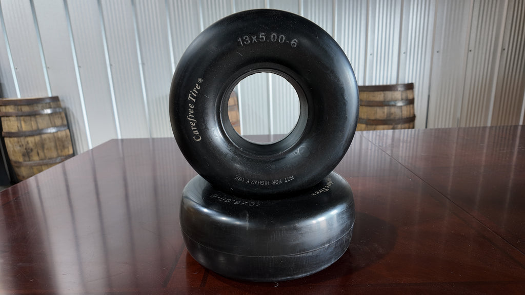 Two black rubber off-road tires stacked on top of each other on a dark wooden table, with a corrugated metal wall and wooden barrels in the background.